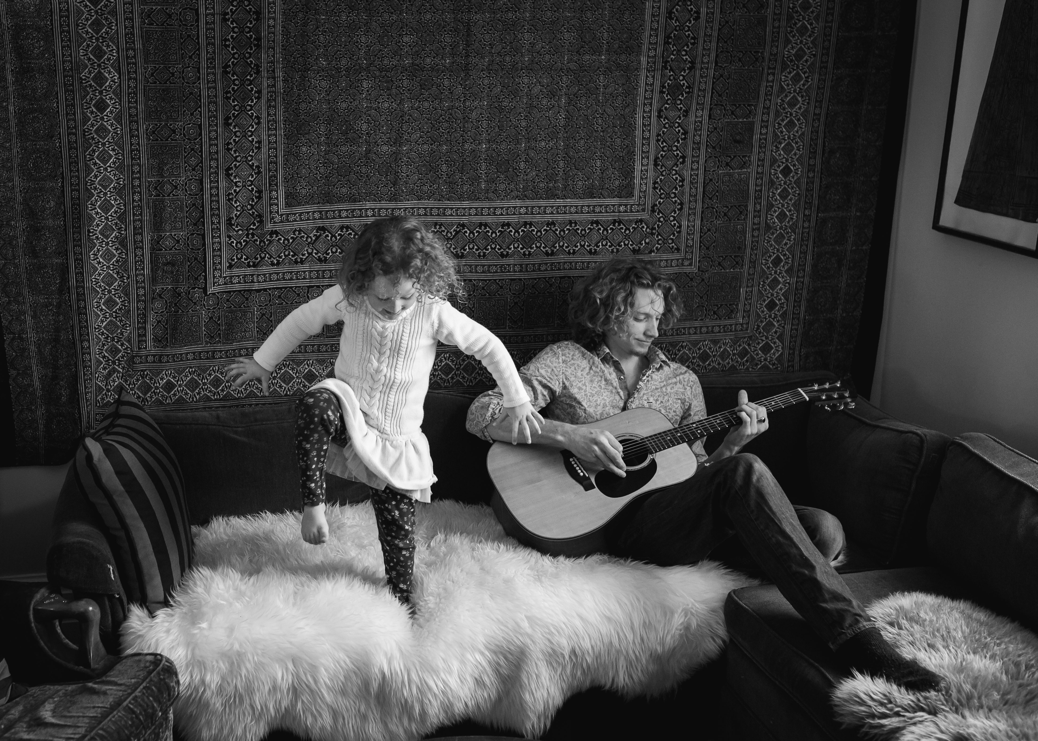 black and white lifestyle image of young girl dancing on couch dad playing guitar