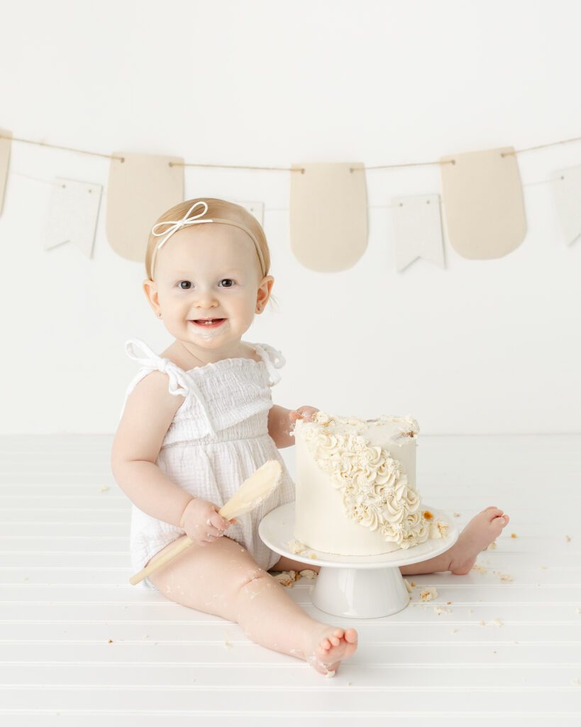 A baby girl in a white romper sits next to a partially smashed cake, gazing at the camera with a mischievous smile. Crumbs are scattered around, adding to the charm of her first birthday portrait.