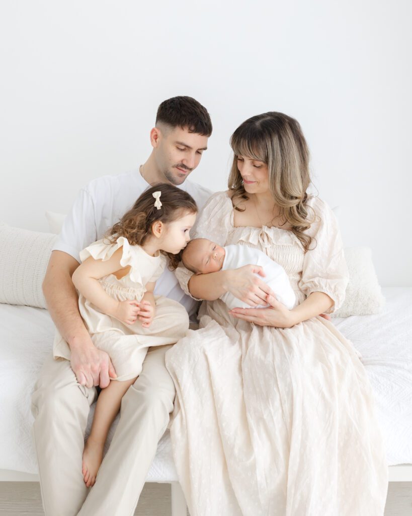 Parents seated on a white bed holding their newborn while the older sibling leans in close, all dressed in light, neutral tones