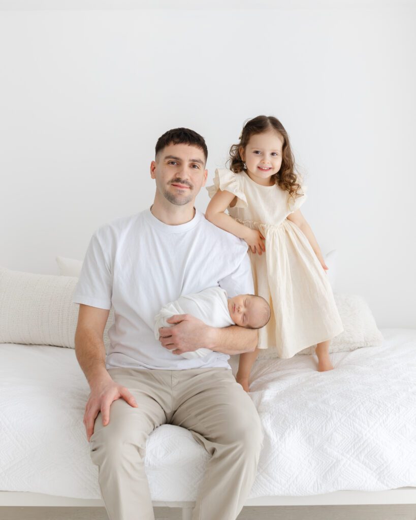 Father sitting on a white bed holding the newborn while the older sibling stands beside him, all in neutral clothing in a bright newborn studio in guelph